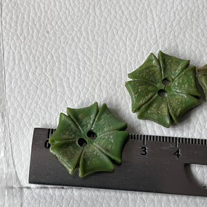 May include: Two olive green buttons with a six-petaled flower shape and a mottled texture. Each button features two central holes for attachment. A black metal ruler is positioned beneath the buttons, showing a width of approximately 2.5 cm.