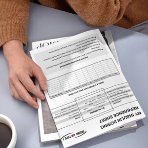 May include: A white reference sheet titled "My Insulin Dosing Reference Sheet" with text and tables, held by a person wearing a brown sweater. A cup of coffee sits nearby. The sheet includes notes and calculations.