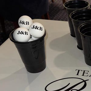 以下が含まれることがあります： 黒いプラスチックカップに白いピンポン玉が詰められ、それぞれに黒で「J&B」と印刷されています。白い表面には他の黒いカップもいくつか見え、ゲームのセットアップを示唆しています。シーンは明るく照らされています。