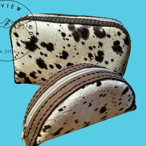 May include: Two cowhide makeup bags with brown leather trim. The larger bag is rectangular and the smaller bag is half-moon shaped. Both bags have a zipper closure.