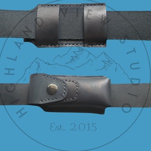 May include: Black leather belt holster with a snap closure. The holster is designed to hold a small, rectangular object, such as a knife or a multi-tool. The holster is attached to a black leather belt. The text "Est. 2015" is visible on the belt.