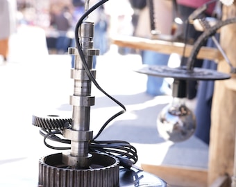 May include: A unique table lamp made from repurposed metal parts, including gears and a camshaft. The lamp has a black cord and a silver light bulb. The lamp is sitting on a gray base.