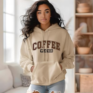 May include: A beige hoodie with a front pocket and a drawstring hood. The word "COFFEE" is printed in brown block letters above the word "WEATHER" in a smaller font. The model is wearing blue jeans.