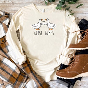 May include: A long-sleeved, cream-coloured t-shirt with a graphic of two geese and the text "Goose Bumps". The shirt is laid out on a wooden surface with a plaid shirt, blue jeans, and brown boots.