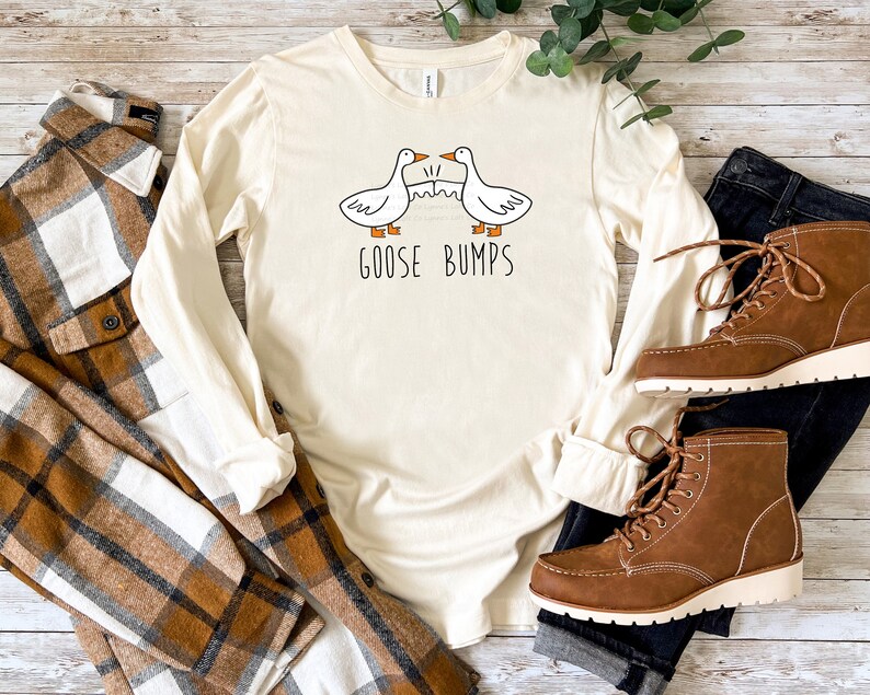 May include: A long-sleeved, cream-coloured t-shirt with a graphic of two geese and the text "Goose Bumps". The shirt is laid out on a wooden surface with a plaid shirt, blue jeans, and brown boots.