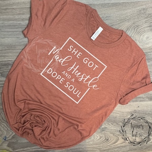 May include: A rust-colored t-shirt with white text that reads "She got Mad Hustle and a Dope Soul". The text is enclosed in a white square.