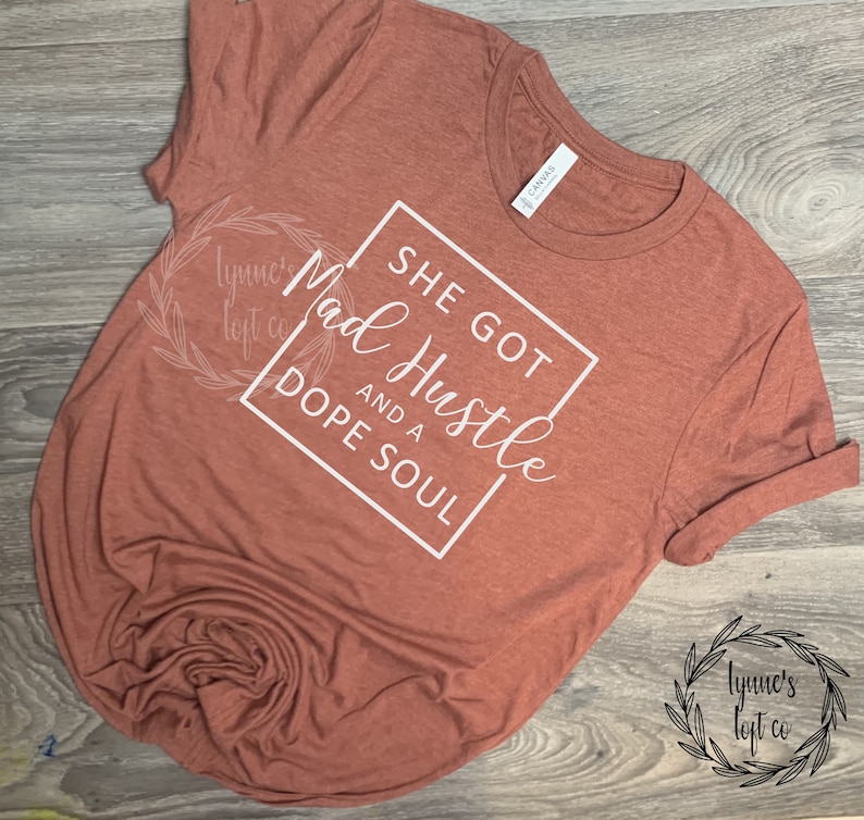 May include: A rust-colored t-shirt with white text that reads "She got Mad Hustle and a Dope Soul". The text is enclosed in a white square.