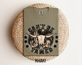 以下が含まれることがあります： カーキ色のTシャツ。牛の頭蓋骨、サボテン、そして「CAN'T BE TAMED」の文字が描かれたグラフィックデザイン。デザインには星と太陽が含まれています。シャツは織られた表面に折りたたまれています。