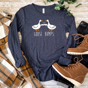 May include: A navy blue long-sleeved t-shirt with a white graphic of two geese facing each other with the text "Goose Bumps". The shirt is laid out on a wooden surface with a plaid flannel shirt, a pair of jeans, and brown boots.