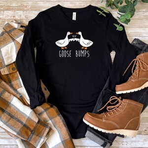 May include: Black long-sleeved shirt with a white graphic of two geese facing each other with the text "Goose Bumps". The shirt is lying on a wooden surface with a brown and white checked shirt and blue jeans with brown boots.