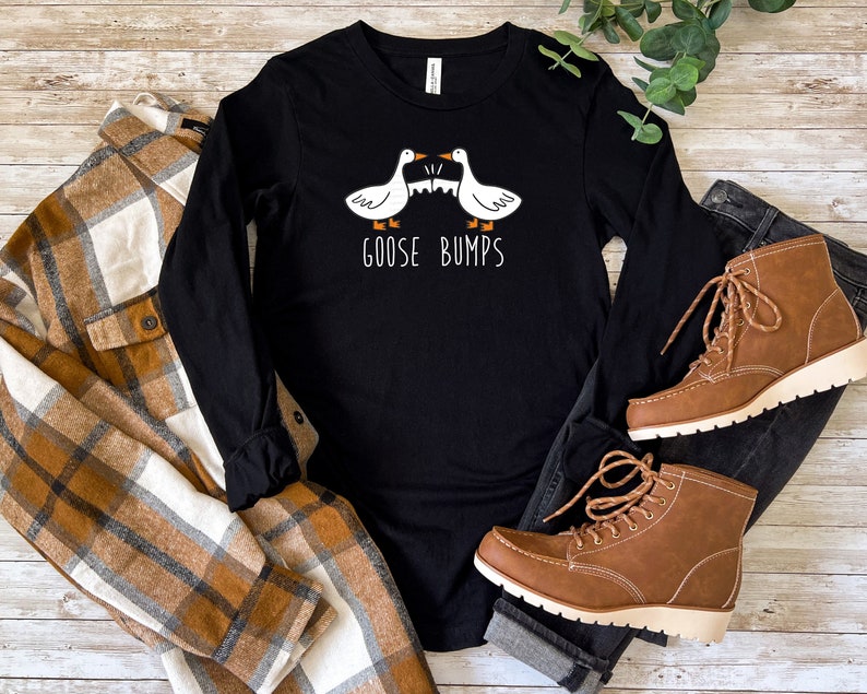 May include: Black long-sleeved shirt with a white graphic of two geese facing each other with the text "Goose Bumps". The shirt is lying on a wooden surface with a brown and white checked shirt and blue jeans with brown boots.