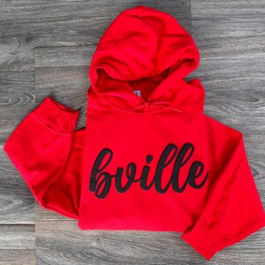 以下が含まれることがあります： 黒い文字で「bville」と書いてある赤いフード付きスウェットシャツ。文字は筆記体です。