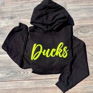 以下が含まれることがあります： 前面に蛍光緑色の「Ducks」のグラフィックがプリントされた黒いフード付きスウェットシャツです。