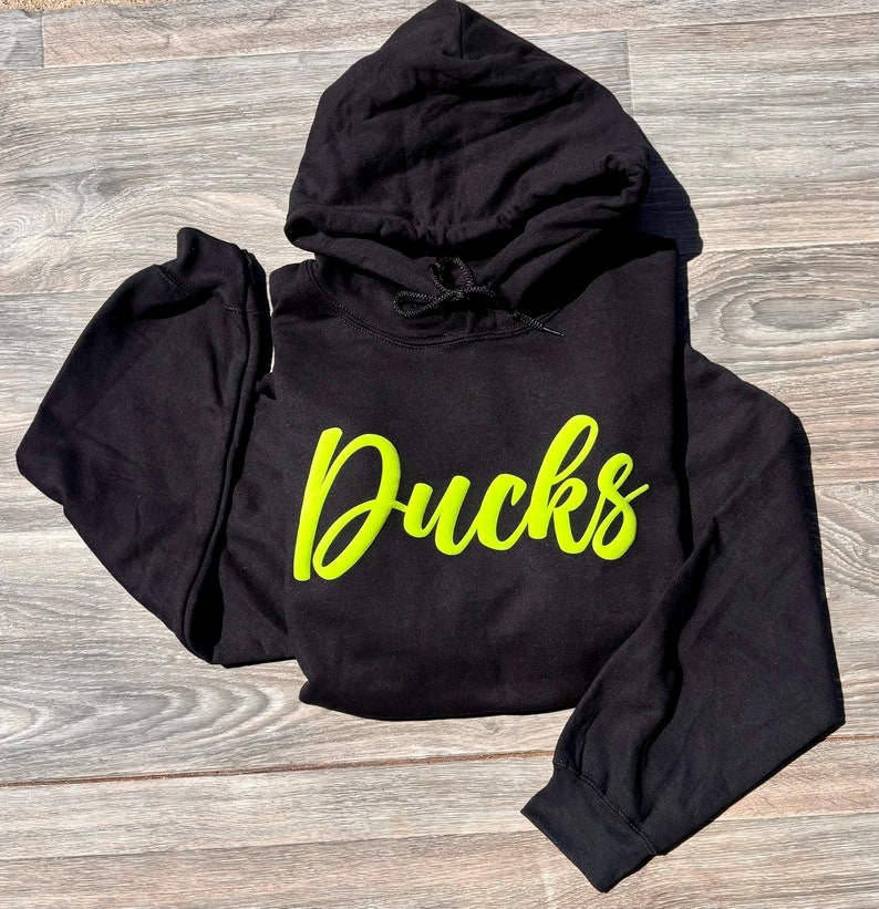 以下が含まれることがあります： 前面に蛍光緑色の「Ducks」のグラフィックがプリントされた黒いフード付きスウェットシャツです。