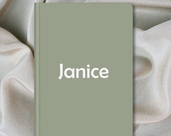 Op de afbeelding: Een saliegroen notitieboek met de naam "Janice" in het wit gedrukt. Het notitieboek is gesloten en heeft een gladde kaft. De achtergrond is een zachte, crèmekleurige stof.
