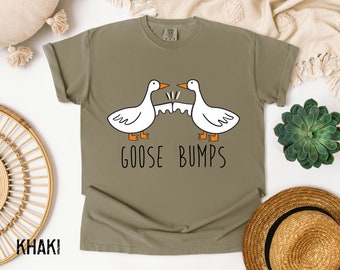 May include: A khaki green t-shirt with a white graphic of two geese bumping into each other. The text "Goose Bumps" is printed below the geese.