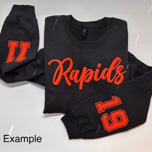 以下が含まれることがあります： 前面に赤い文字で「Rapids」と書かれた黒いスウェットシャツ。片方の袖には赤い数字「11」、もう片方の袖には赤い数字「16」が書かれている。