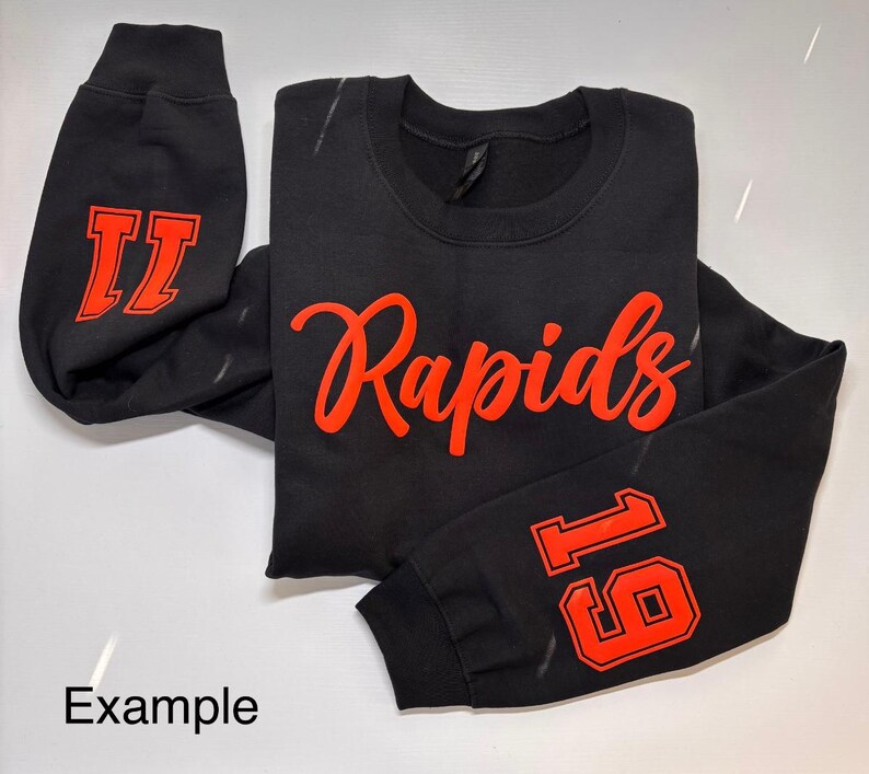 以下が含まれることがあります： 前面に赤い文字で「Rapids」と書かれた黒いスウェットシャツ。片方の袖には赤い数字「11」、もう片方の袖には赤い数字「16」が書かれている。
