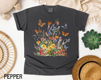 May include: A dark grey t-shirt with a watercolour design of wildflowers and butterflies. The design features orange, blue, yellow, and red flowers, with several butterflies. The word "PEPPER" is printed at the bottom of the shirt.