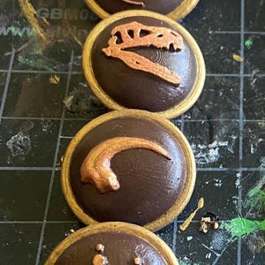 May include: Four brown, circular buttons with gold rims and bronze-colored dinosaur-themed designs. The designs include a dragon, a skull, a claw, and a footprint. The buttons are arranged vertically.