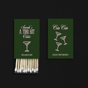 May include: Green matchbox with the text "Sarah's A Tini Bit Older" and a martini glass illustration. The matchbox is open, revealing the matchsticks. The adjacent matchbox reads "Cin Cin" with martini glasses.