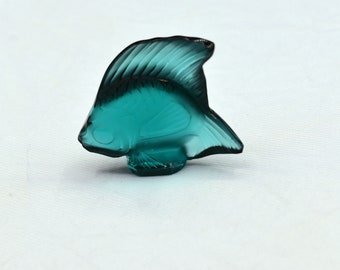 May include: A teal-colored glass fish figurine. The fish has detailed fins and a textured body. The figurine is displayed on a white surface. This decorative item could be used as a paperweight or a unique home decor accent.