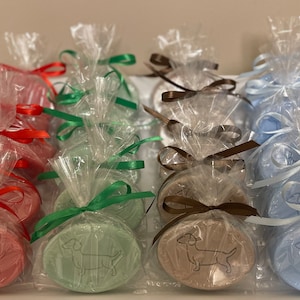 Op de afbeelding: Ovale zepen in verschillende kleuren, elk verpakt in transparant cellofaan en vastgebonden met een lint. De zepen hebben een teckel-hond ontwerp. Kleuren zijn rood, groen, bruin en blauw. Gepresenteerd in een witte tray.