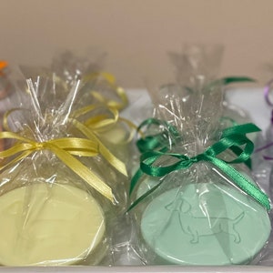 Op de afbeelding: Ovale zepen in pastelkleuren, elk met een honden silhouet, zijn verpakt in doorzichtig cellofaan en vastgebonden met kleurrijke linten. De zepen zijn in tinten van oranje, geel, groen en paars.