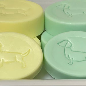 Op de afbeelding: Een verzameling pastelkleurige, ronde zepen. Elke zeep heeft een reli&euml;fdesign van een teckel. Kleuren zijn onder meer perzik, geel, mintgroen en lavendel. De zepen zijn gestapeld in een witte container.