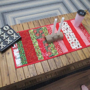 Puede incluir: Un camino de mesa de Navidad rojo y verde con una variedad de patrones, incluyendo copos de nieve, bastones de caramelo y árboles de Navidad. El camino de mesa está sobre una mesa de madera con dos velas blancas y un porta-troncos de madera.