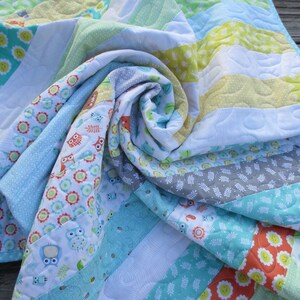 May include: A colorful, quilted blanket with a variety of patterns and colors. The blanket features stripes of white, blue, green, yellow, and orange fabrics. Some stripes have floral and owl patterns. The blanket is made of soft fabric and has a quilted design.