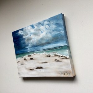 May include: Oil painting of a beach scene with a cloudy sky. The artwork depicts a white sandy beach with small rocks, turquoise water, and a dramatic sky with shades of blue and grey. The painting is on a square canvas.