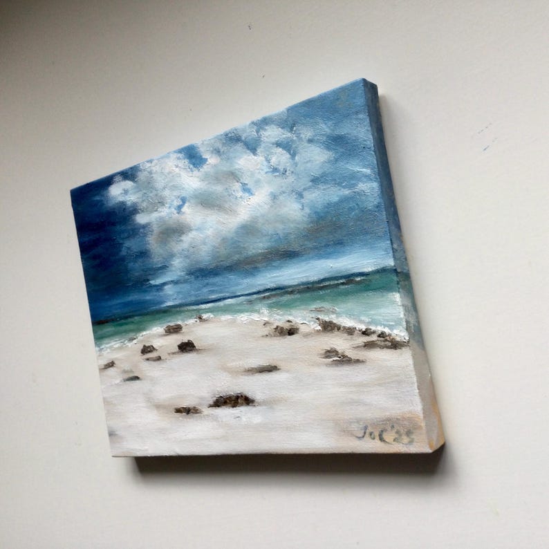 May include: Oil painting of a beach scene with a cloudy sky. The artwork depicts a white sandy beach with small rocks, turquoise water, and a dramatic sky with shades of blue and grey. The painting is on a square canvas.