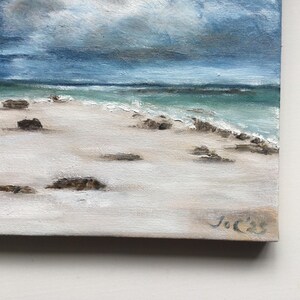 May include: Oil painting depicting a beach scene with a white sandy shore, turquoise water, and a cloudy sky. Dark rocks are scattered on the sand. The painting is signed in the bottom right corner.