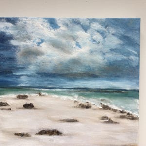May include: An oil painting of a beach scene under a cloudy sky. The artwork shows a white sandy beach, turquoise water, and dark, textured rocks. The sky is filled with shades of blue and grey, creating a dramatic atmosphere.
