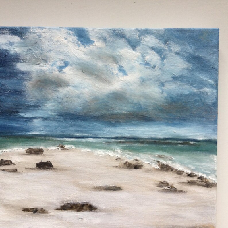 May include: An oil painting of a beach scene under a cloudy sky. The artwork shows a white sandy beach, turquoise water, and dark, textured rocks. The sky is filled with shades of blue and grey, creating a dramatic atmosphere.