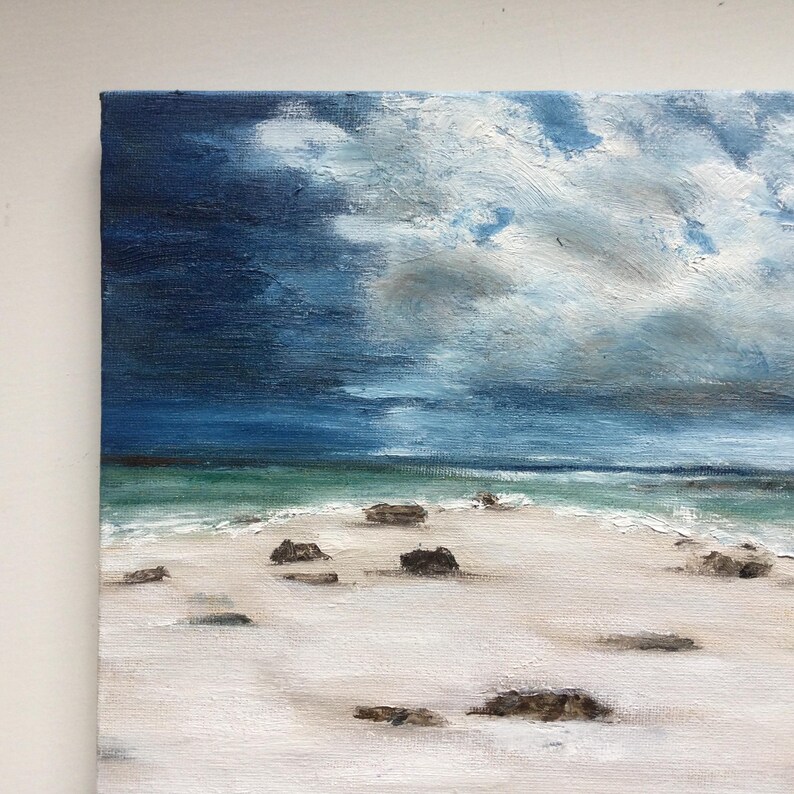 May include: An oil painting of a beach scene under a dramatic sky. The sky transitions from dark blue to lighter shades, with fluffy white clouds. The beach has white sand and scattered dark rocks, with turquoise water in the distance.
