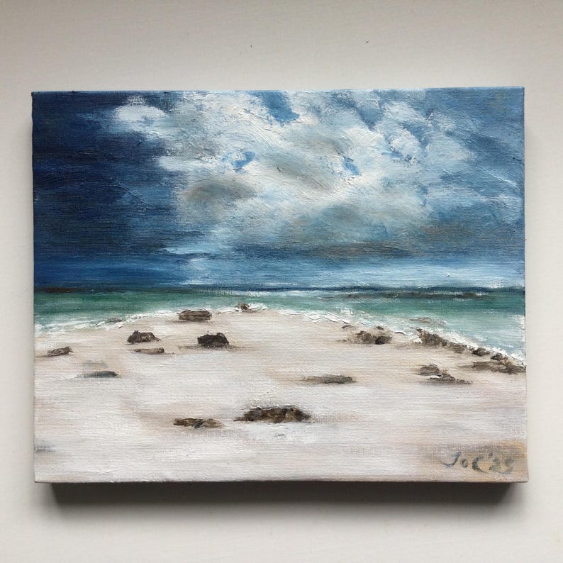 May include: Oil painting of a beach scene with a stormy sky. The artwork features a white sandy beach with scattered rocks, turquoise water, and a dark blue, cloudy sky. The artist's signature is in the bottom right corner.