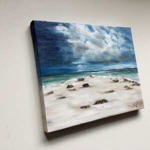 May include: Square oil painting depicting a beach scene under a cloudy sky. The artwork shows a white sandy beach, turquoise water, and dark rocks. The sky is a blend of dark blue and grey, with white clouds. The artist's signature is in the bottom right corner.