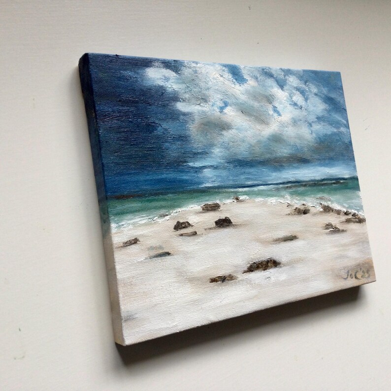 May include: Square oil painting depicting a beach scene under a cloudy sky. The artwork shows a white sandy beach, turquoise water, and dark rocks. The sky is a blend of dark blue and grey, with white clouds. The artist's signature is in the bottom right corner.