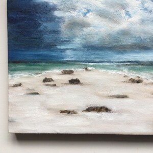 May include: An oil painting of a beach scene under a stormy sky. The artwork shows a white sandy beach with dark rocks, turquoise water, and a dramatic sky with shades of blue and grey.