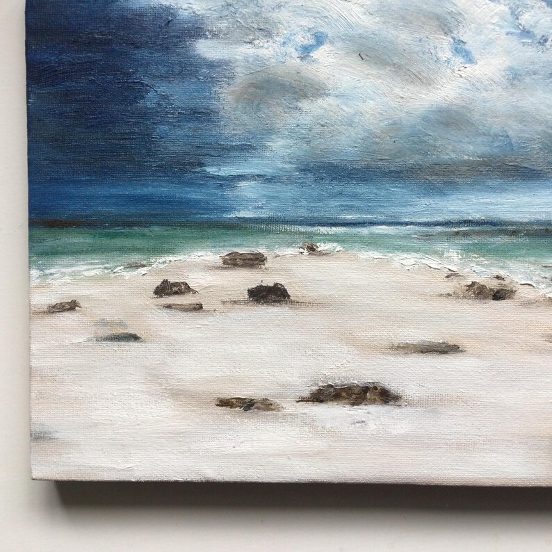 May include: An oil painting of a beach scene under a stormy sky. The artwork shows a white sandy beach with dark rocks, turquoise water, and a dramatic sky with shades of blue and grey.