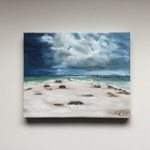 May include: Oil painting depicting a beach scene under a dramatic sky. The artwork shows a white sandy beach, turquoise water, and dark blue clouds. Small rocks are on the sand. The painting is signed in the bottom right corner.