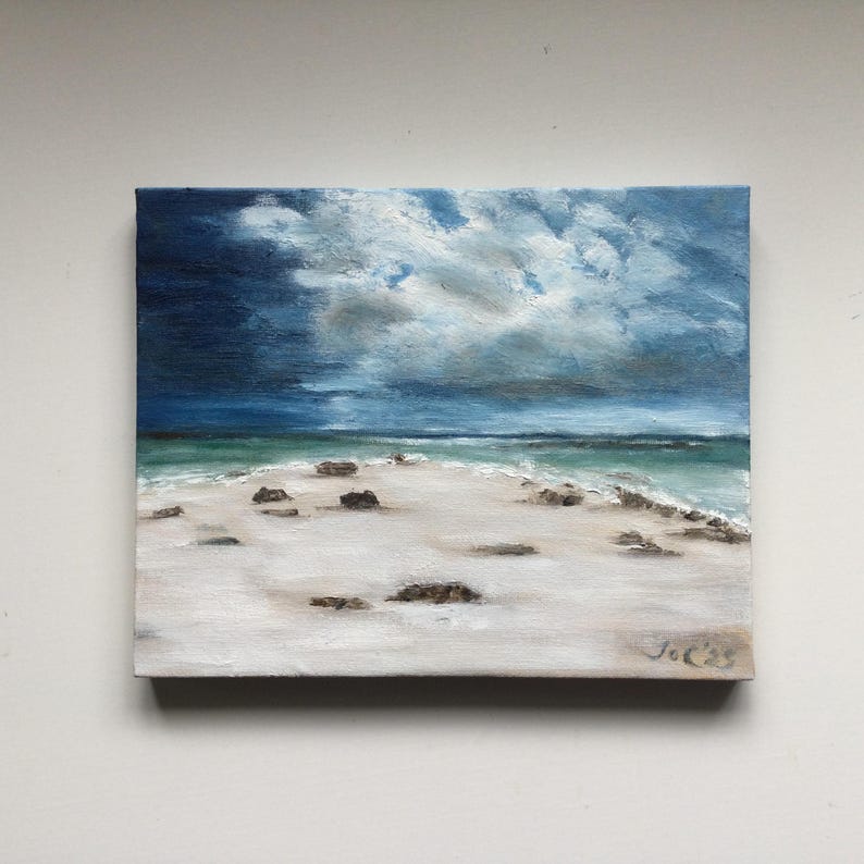 May include: Oil painting depicting a beach scene under a dramatic sky. The artwork shows a white sandy beach, turquoise water, and dark blue clouds. Small rocks are on the sand. The painting is signed in the bottom right corner.