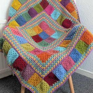 May include: A colorful, handmade crochet blanket draped over a white chair. The blanket features a patchwork design with squares in various colors, including red, yellow, blue, and green. The edges are trimmed with a contrasting border.