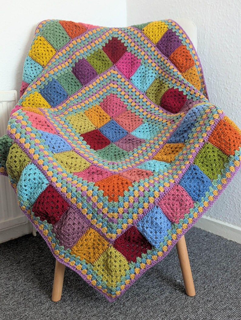 May include: A colorful, handmade crochet blanket draped over a white chair. The blanket features a patchwork design with squares in various colors, including red, yellow, blue, and green. The edges are trimmed with a contrasting border.