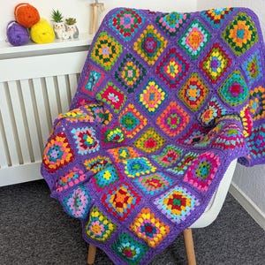 May include: A handmade, crocheted blanket draped over a white chair. The blanket features a vibrant patchwork design of colorful squares in shades of red, orange, yellow, green, blue, and pink, set against a purple background. The blanket is made of soft yarn.