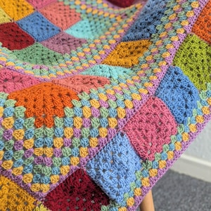 May include: A colorful, handmade crochet blanket with a patchwork design. The blanket features squares in various colors, including pink, blue, orange, and green. The blanket is draped over a wooden chair, showcasing its texture and vibrant hues.
