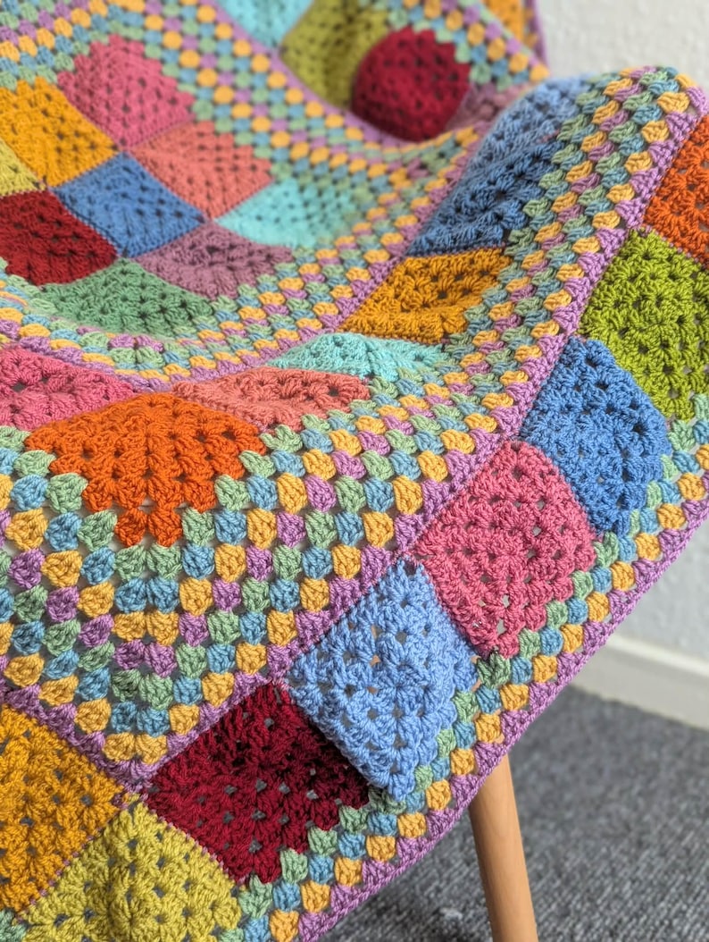 May include: A colorful, handmade crochet blanket with a patchwork design. The blanket features squares in various colors, including pink, blue, orange, and green. The blanket is draped over a wooden chair, showcasing its texture and vibrant hues.