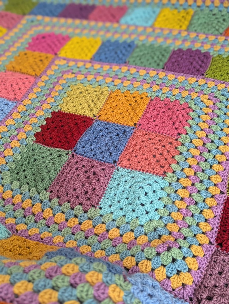 May include: A handmade, colorful crochet blanket featuring a patchwork design. The blanket showcases squares in various shades, including pink, yellow, blue, green, and purple. The squares are bordered by a light blue and orange trim, creating a vibrant and cozy textile.